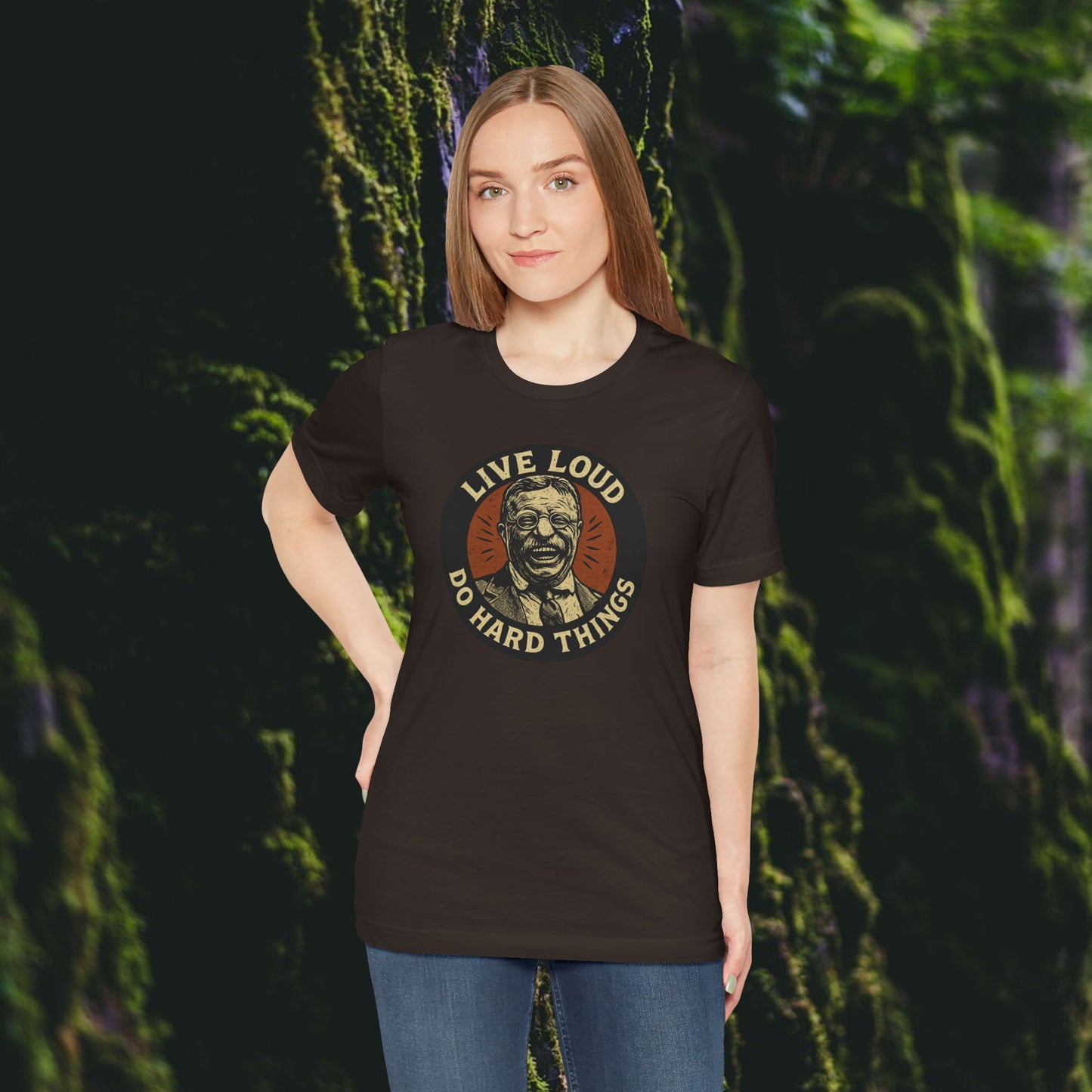 A woman with straight brown hair wears the Printify Live Loud, Do Hard Things Teddy Roosevelt Tee, featuring a face graphic, while standing among moss-covered trees in a peaceful forest.