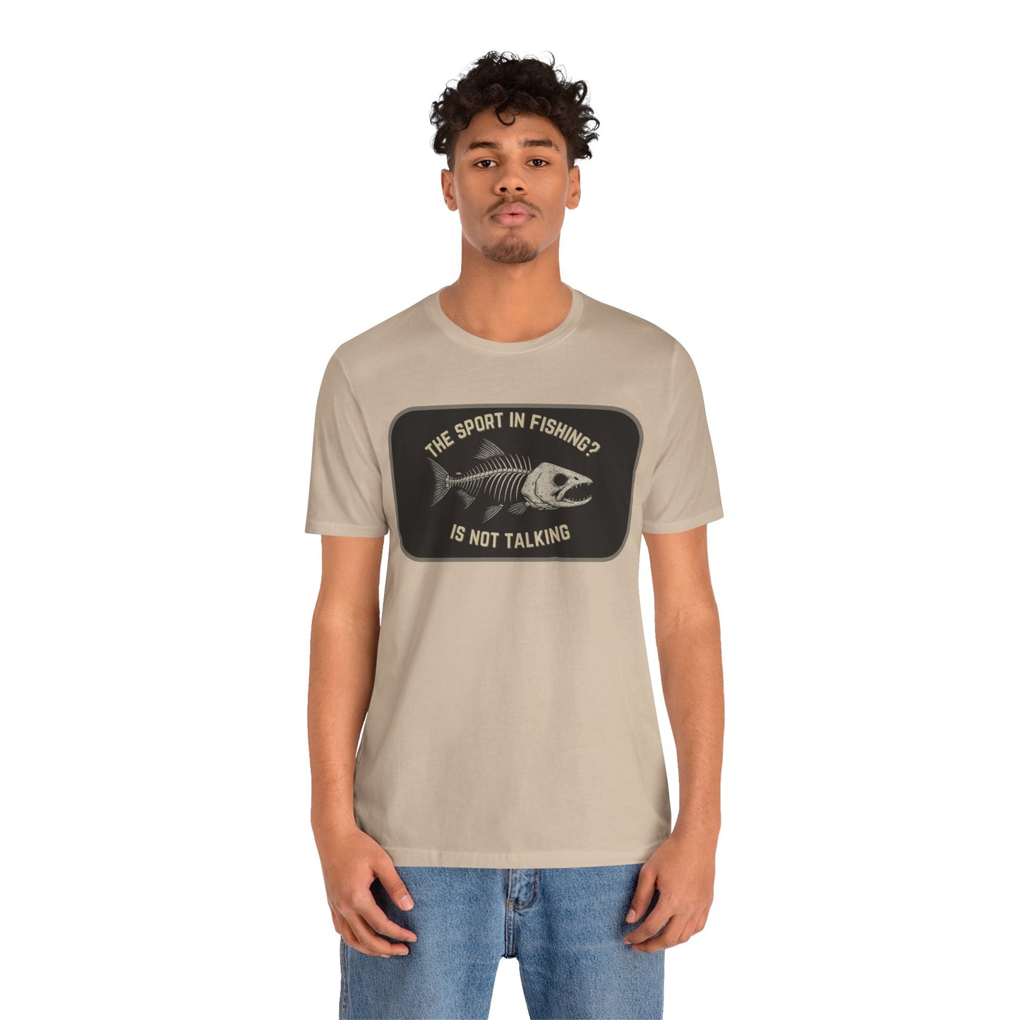 A young man with short curly hair models Printify’s “The Sport in Fishing?” Tee, a beige unisex shirt with a distressed fish skeleton print and slogan, against a plain white background as he looks at the camera.