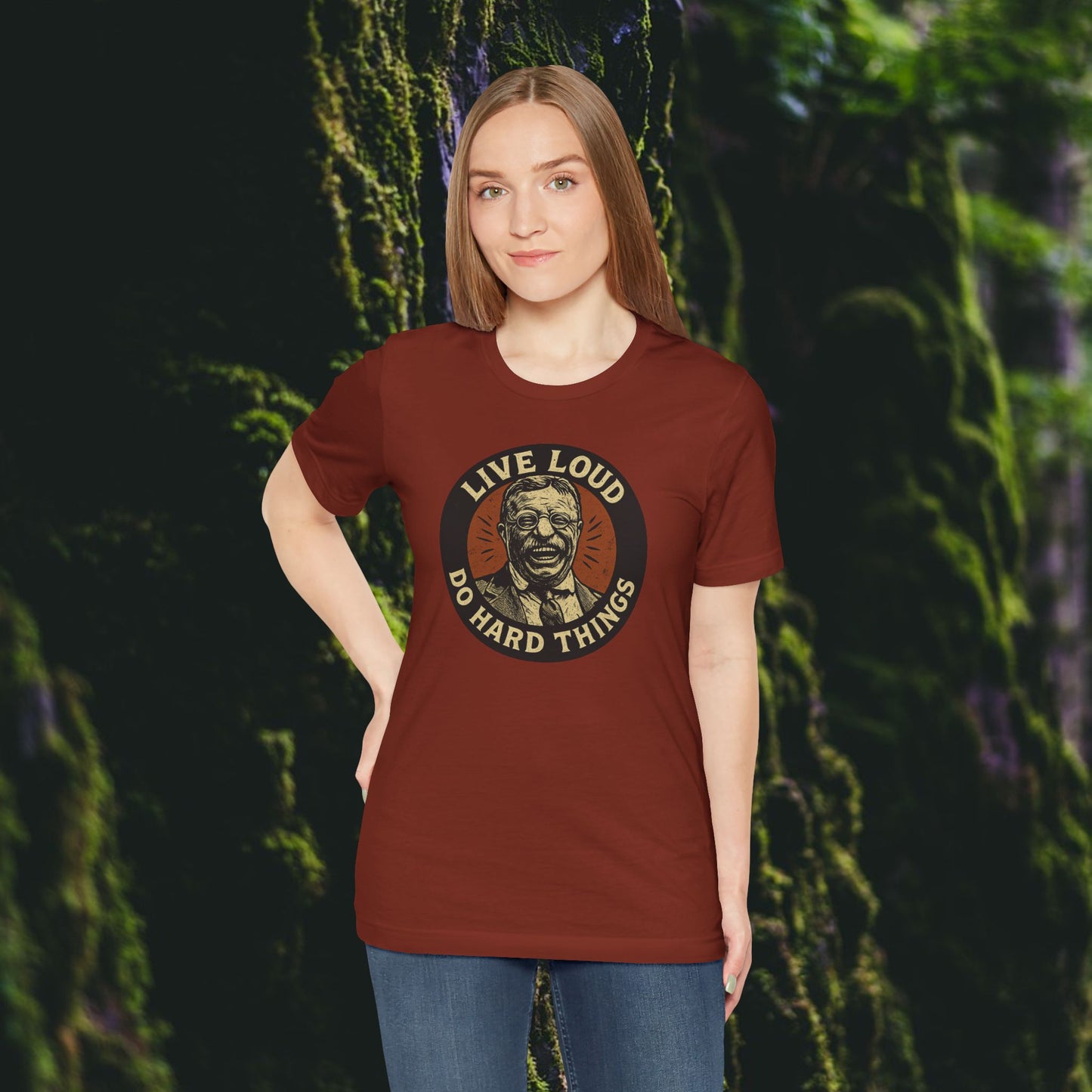 A woman stands before a mossy forest, wearing the Printify Live Loud, Do Hard Things Teddy Roosevelt Tee in maroon, featuring an illustrated face and bold lettering.