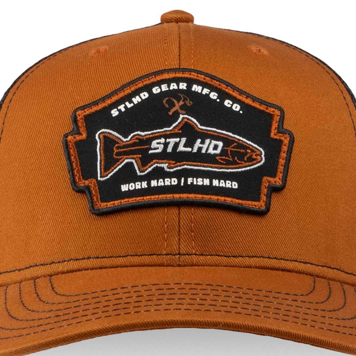 The STLHD Hook X Hammer Trucker Hat by STLHD GEAR is brown with a black and orange patch showing a fish outline and text: STLHD Gear Mfg. Co., STLHD, Work Hard | Fish Hard. It features an adjustable snapback for comfort.