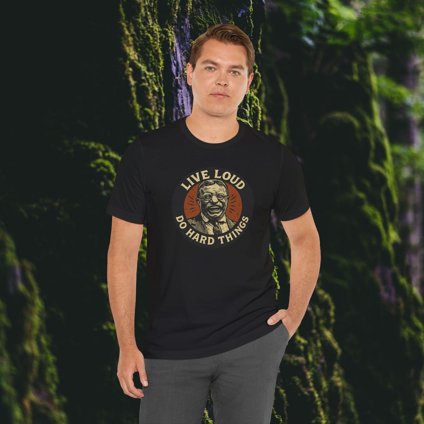 A man wearing the Printify Live Loud, Do Hard Things Teddy Roosevelt Tee stands in front of a mossy forest, one hand in his pocket, reflecting the bold spirit of this inspirational shirt.