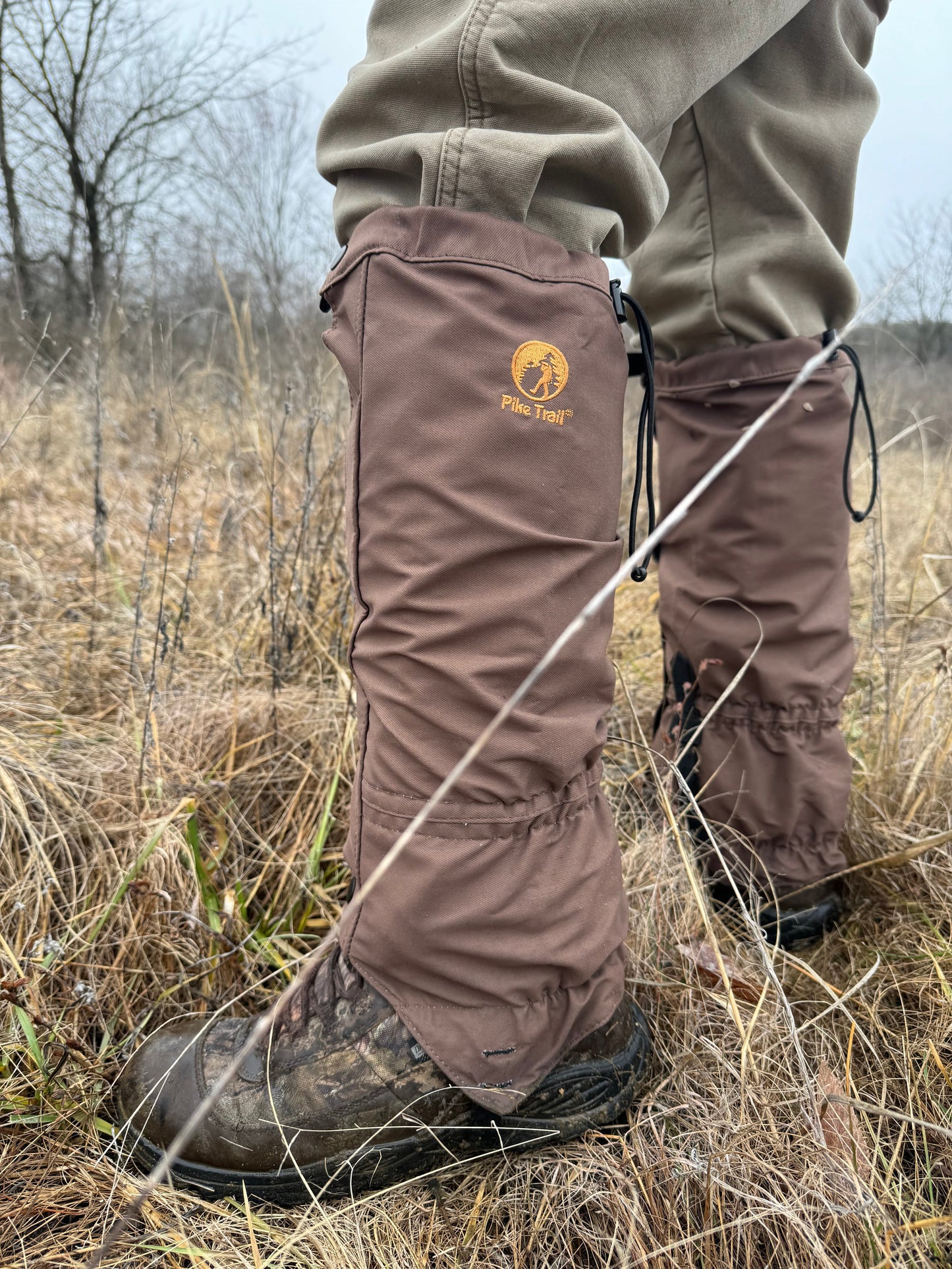 Wearing Pike Trail All-Season Waterproof Leg Gaiters over boots and pants, a person stands in tall grass. These abrasion-resistant gaiters, secured with cords and featuring the Pike Trail logo, protect lower legs in the grassy field.