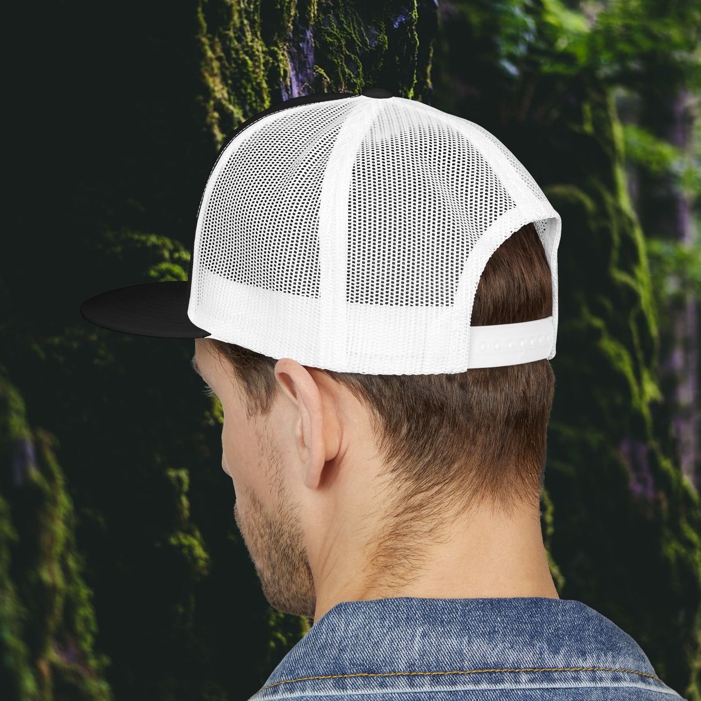 A person seen from behind wears the Printify Public Land Trucker Hat, Not Yours To Sell The Land Resists, featuring white mesh and a black brim, set against a blurred green nature background.