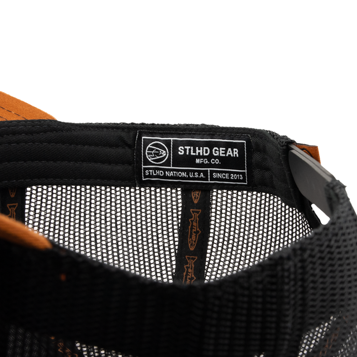 Close-up of the inside of a black mesh STLHD Hook X Hammer Trucker Hat by STLHD GEAR, featuring a label that reads STLHD GEAR MFG. CO. and STLHD NATION. U.S.A. SINCE 2013 with a fish logo on this adjustable snapback hat.