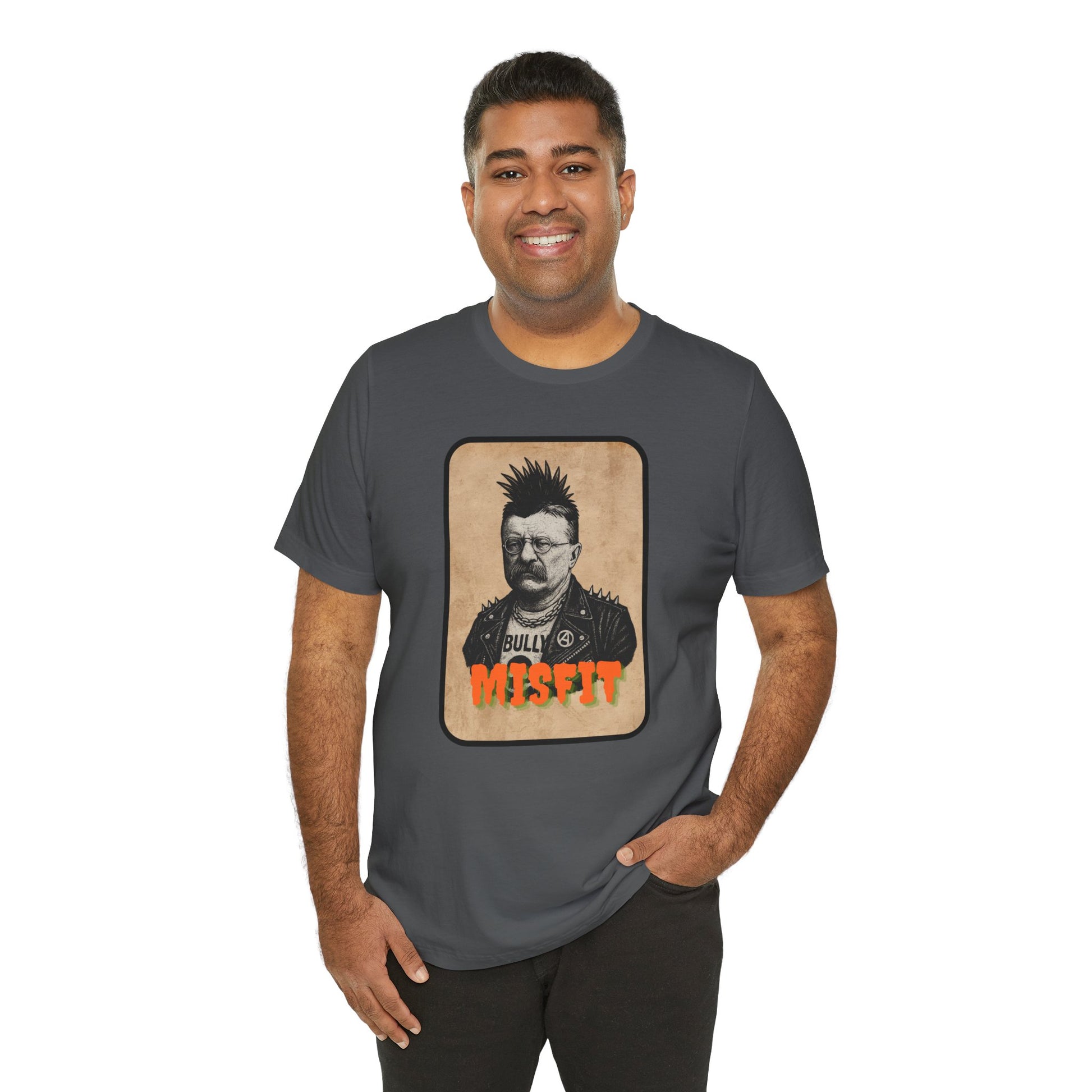 A smiling man wears the Iron Pines Teddy Roosevelt Punk T-Shirt, a dark gray Misfit History tee featuring a vintage-style BULLY graphic and bold orange MISFIT lettering. He stands with one hand in his pocket.