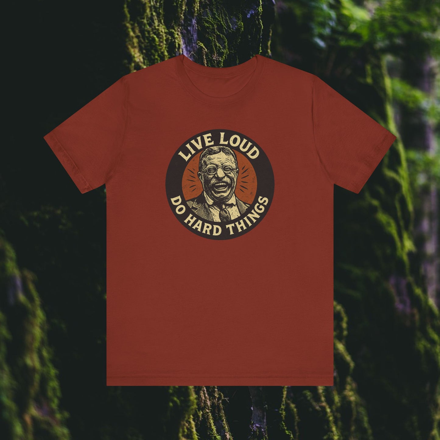 The Live Loud, Do Hard Things Teddy Roosevelt Tee by Printify features a maroon shirt with an illustrated smiling Teddy Roosevelt, encircled by LIVE LOUD and DO HARD THINGS, set on a blurred mossy forest background.
