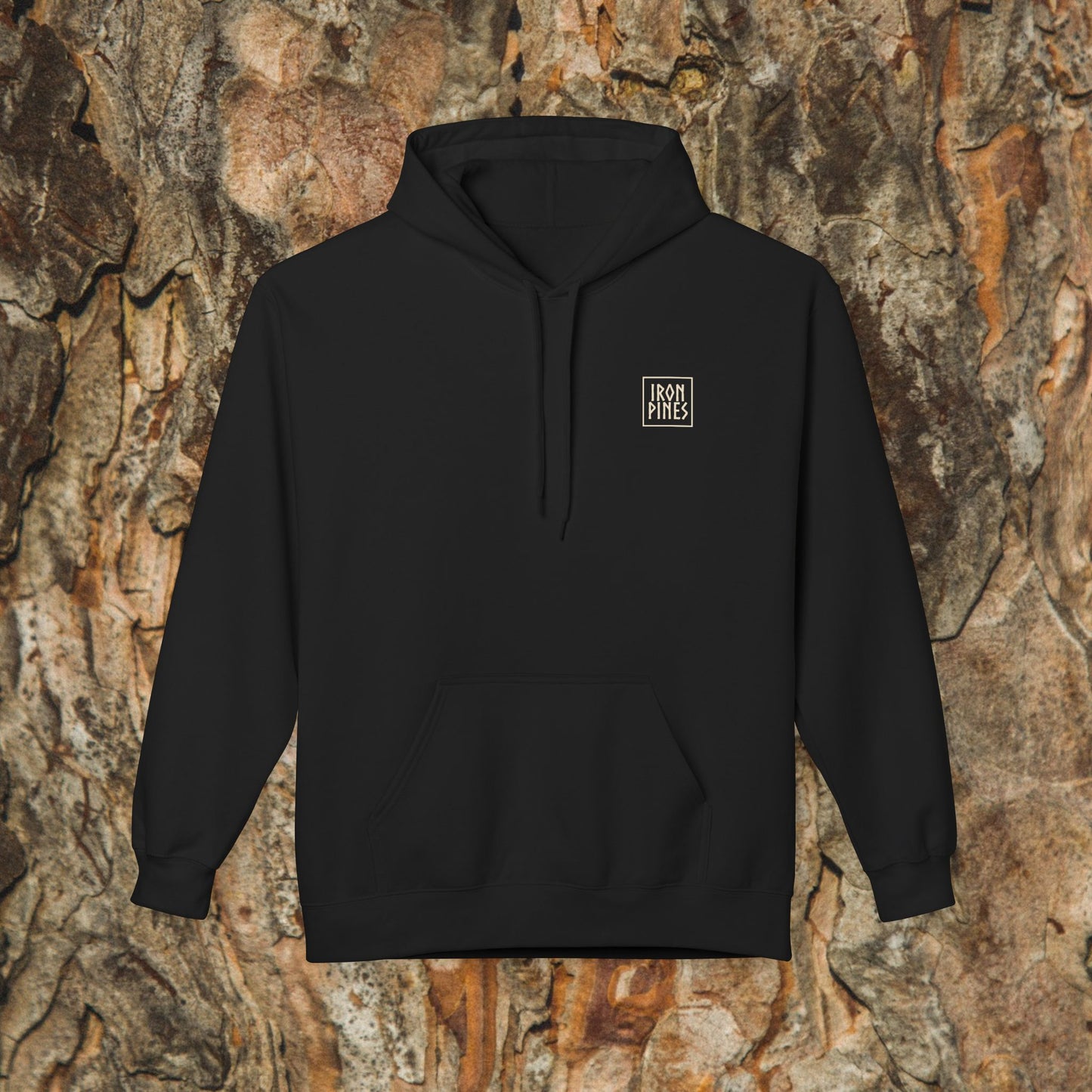 The Iron Pines Hoodie by Printify is a black midweight fleece with a small left-chest logo, shown against tree bark—a sustainable outdoor essential for any adventure.
