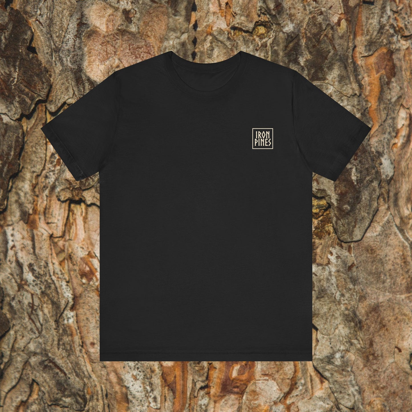 The Iron Pines Crest Tee by Printify is a black, ultra-soft Airlume cotton shirt featuring a small white boxed logo on the upper left chest, shown laid flat against rough tree bark.