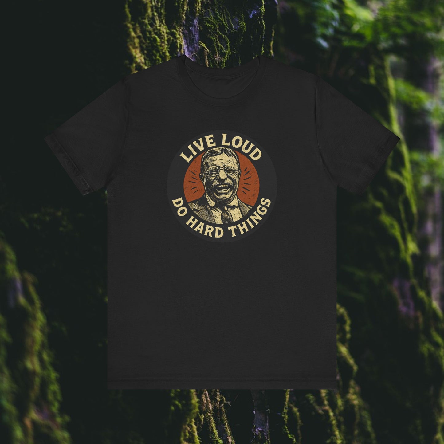 The Live Loud, Do Hard Things Teddy Roosevelt Tee by Printify is a black shirt featuring a circular graphic of a smiling man, motivational text, and a mossy forest backdrop—perfect for bold, inspirational style enthusiasts.