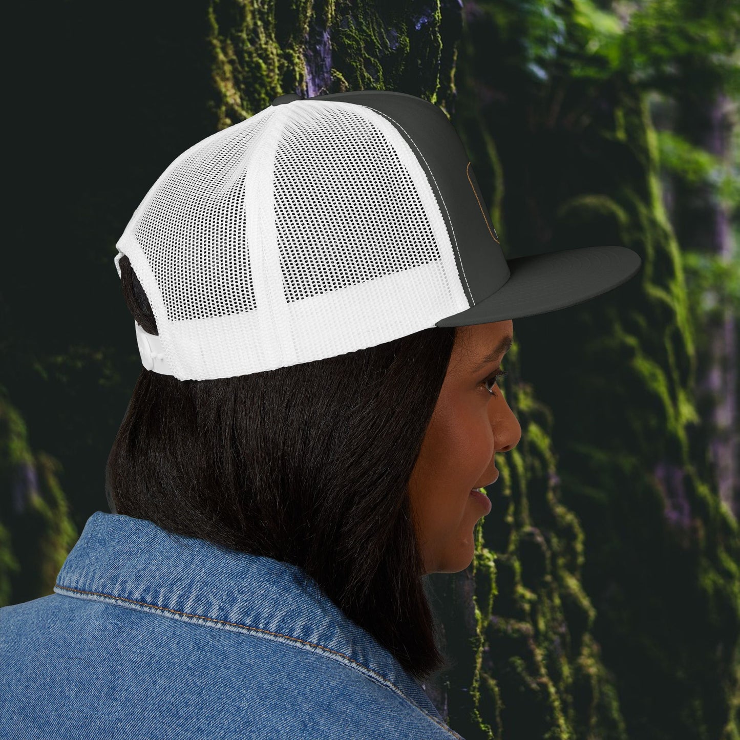 A person in profile wears a Printify Iron Pines Trucker Hat – “Live Loud. Be Inconvenient.” Brass Knuckle Edition, with white and black mesh, and a blue denim jacket, standing by green moss-covered rocks.