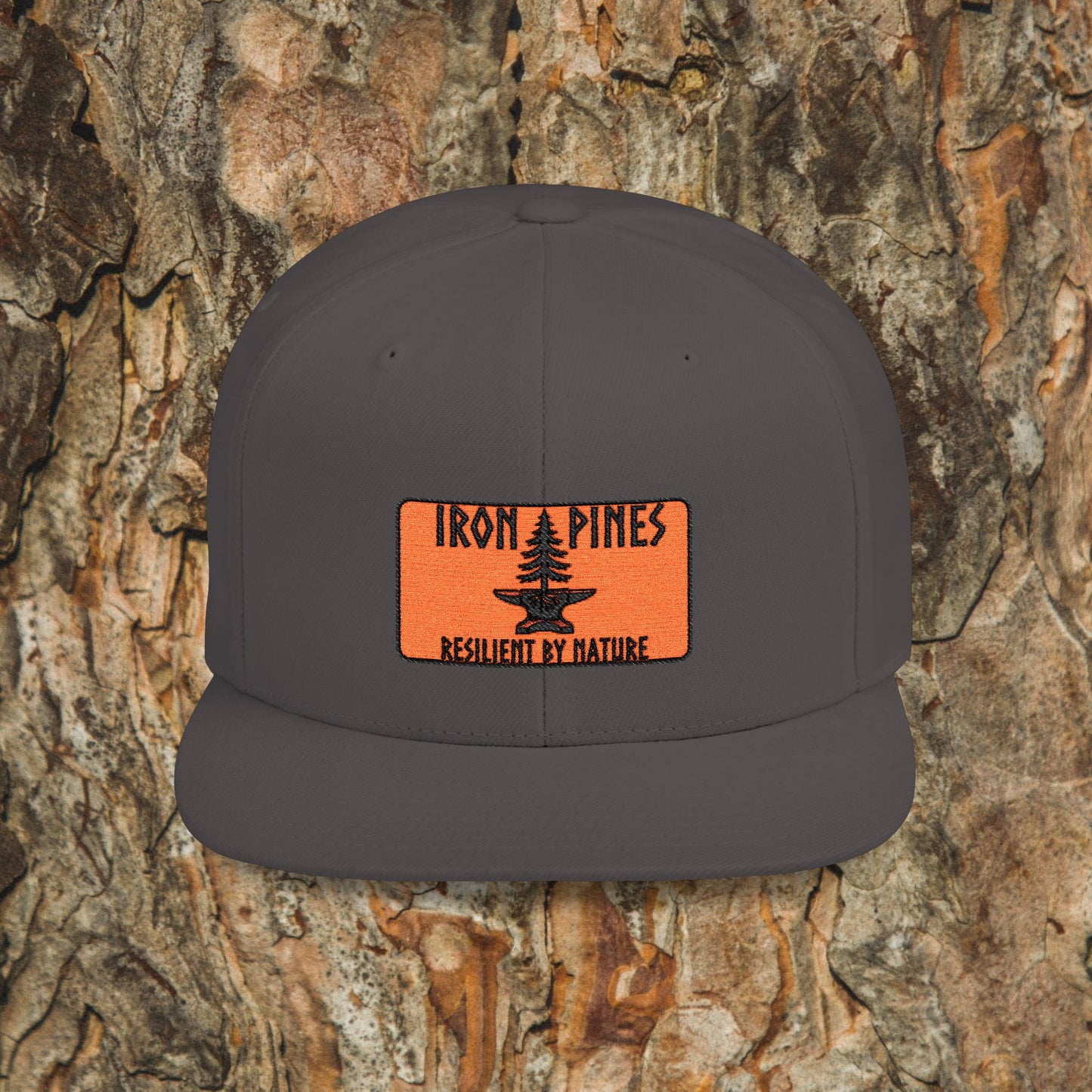 The Iron Pines Logo Hat by Printify features a dark gray design, an orange IRON PINES patch with RESILIENT BY NATURE above a black pine tree graphic, and is shown as a structured snapback against textured tree bark.