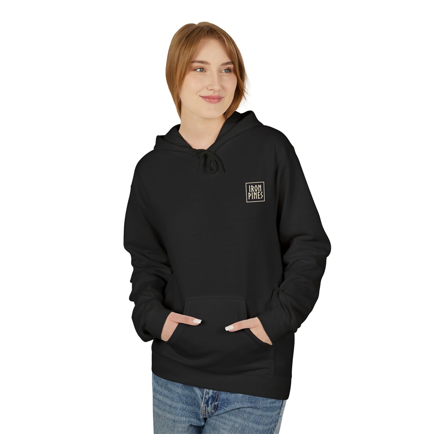 A person with light brown hair wears The Iron Pines Hoodie by Printify, a sustainable black outdoor hoodie with a small white chest logo. Smiling and looking to the side, they keep their hands in the front pocket.