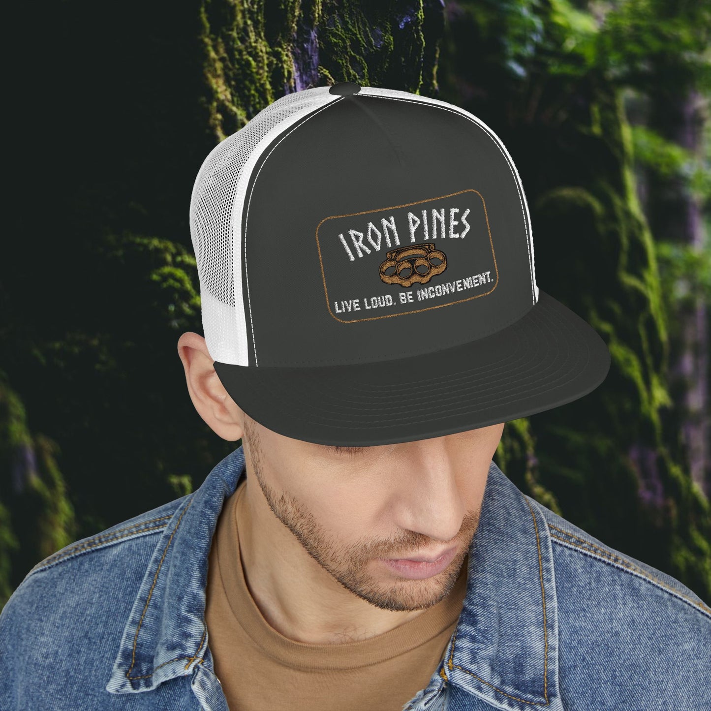 A man in a denim jacket looks down against a forest backdrop, wearing the Printify Iron Pines Trucker Hat – “Live Loud. Be Inconvenient.” Brass Knuckle Edition, featuring a classic black and white mesh snapback style with statement patch.