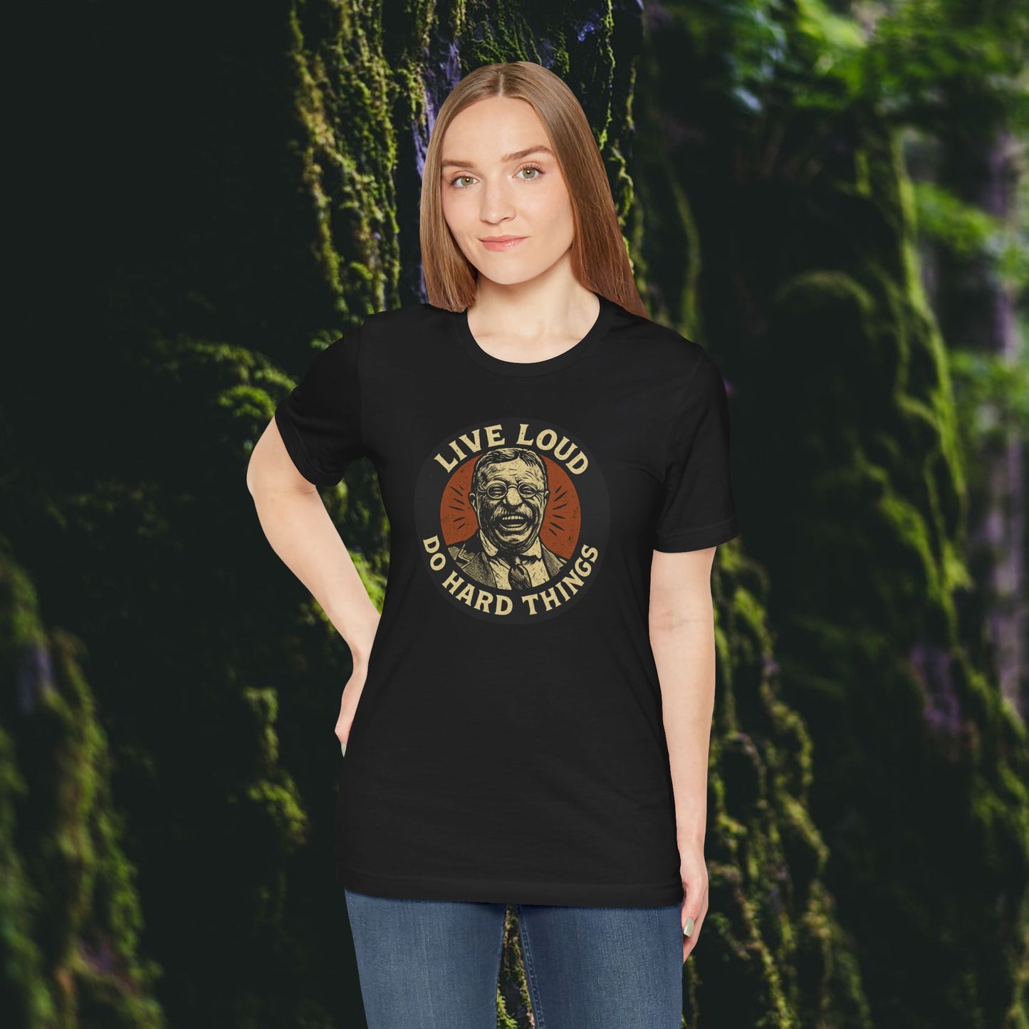 A woman with straight brown hair stands before a lush mossy backdrop, wearing the Printify Live Loud, Do Hard Things Teddy Roosevelt Tee featuring an illustrated portrait of Roosevelt at the center.