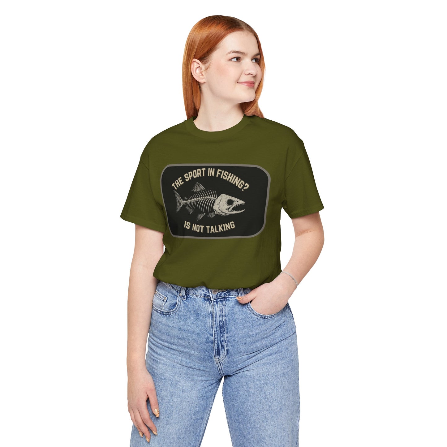 A woman with long red hair smiles while wearing Printify’s “The Sport in Fishing?” Tee, a green shirt with a distressed fish skeleton and the phrase THE SPORT IN FISHING? IS NOT TALKING. She stands with one hand in her jeans pocket, looking to the side.