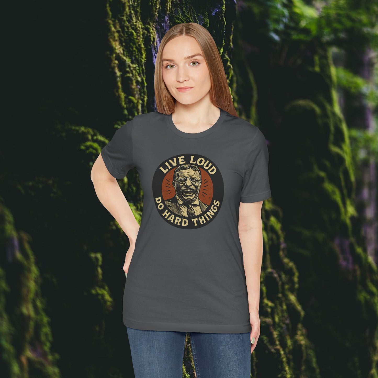 A woman with straight brown hair stands before a mossy rock wall, smiling slightly with one hand on her hip, wearing the Printify Live Loud, Do Hard Things Teddy Roosevelt Tee and radiating the spirit of inspiration.