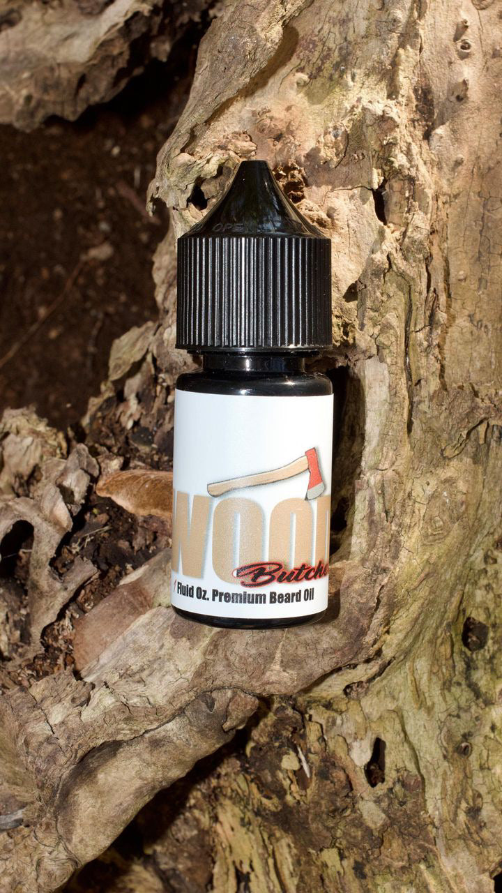 A bottle of Bedlam Beard Companys Wood Butcher beard oil—featuring mahogany, teakwood, vanilla, oak, amber, and musk—rests in a hollow section of textured tree bark.
