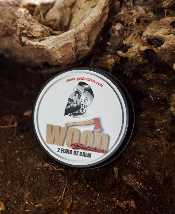 A round tin labeled Wood Butcher by Bedlam Beard Company—featuring mahogany, teakwood, vanilla, oak, amber, and musk—rests on soil near rough tree bark; ideal for fans of rugged conditioning beard balm with a woodsy scent.