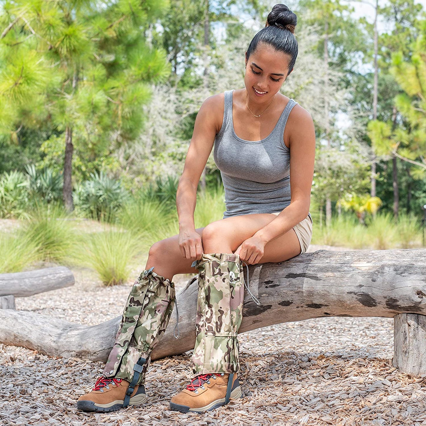 A woman in a gray tank top and shorts sits on a log outdoors, smiling as she puts on Pike Trail All-Season Waterproof Leg Gaiters—built for the trail, the hunt, and everything in between—over her hiking boots amid lush greenery.