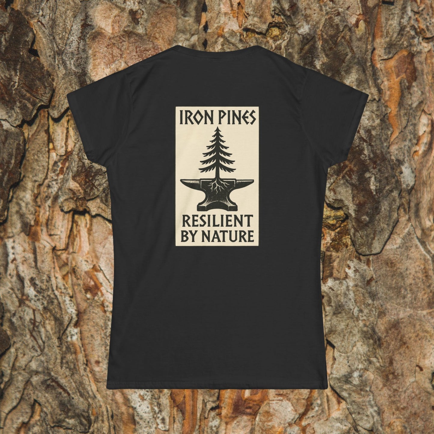 The Iron Pines Woman’s Tee by Printify is a black softstyle shirt featuring a beige pine tree and anvil graphic with “IRON PINES RESILIENT BY NATURE” on the back, shown against tree bark.