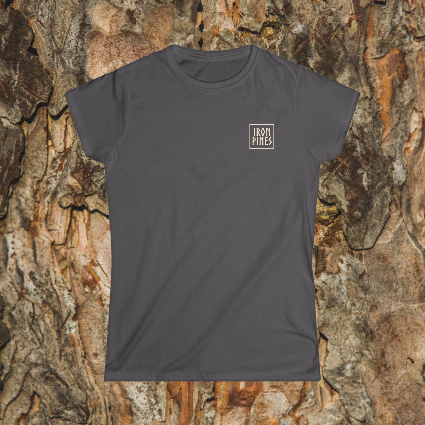 The Iron Pines Womans Tee by Printify in charcoal gray features a small white IRON PINES logo on the left chest, displayed against a textured tree bark background—resilient by nature.