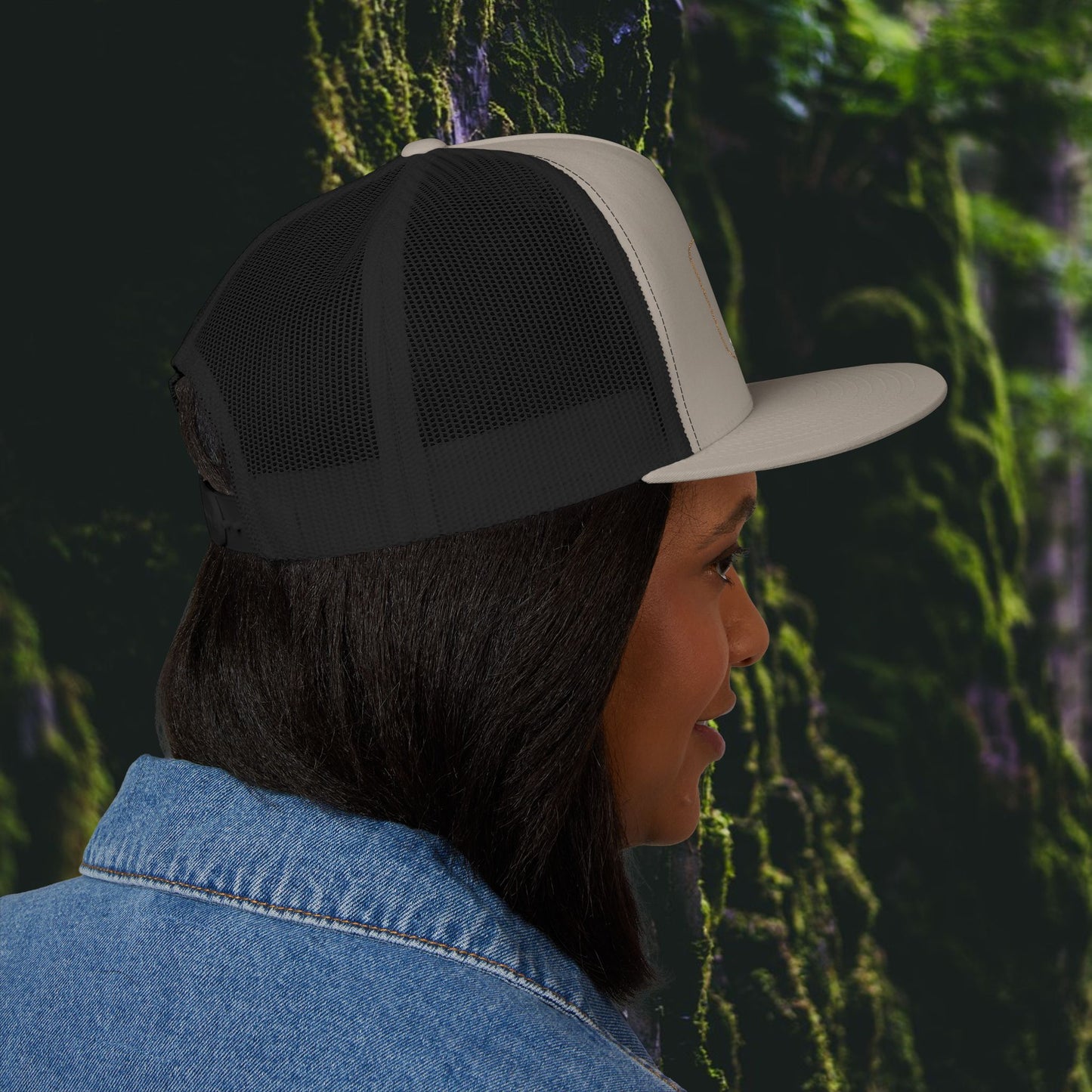A woman with straight dark hair wears the Printify Iron Pines Trucker Hat – “Live Loud. Be Inconvenient.” Brass Knuckle Edition and a blue denim jacket, shown in profile against a lush, green forest background.