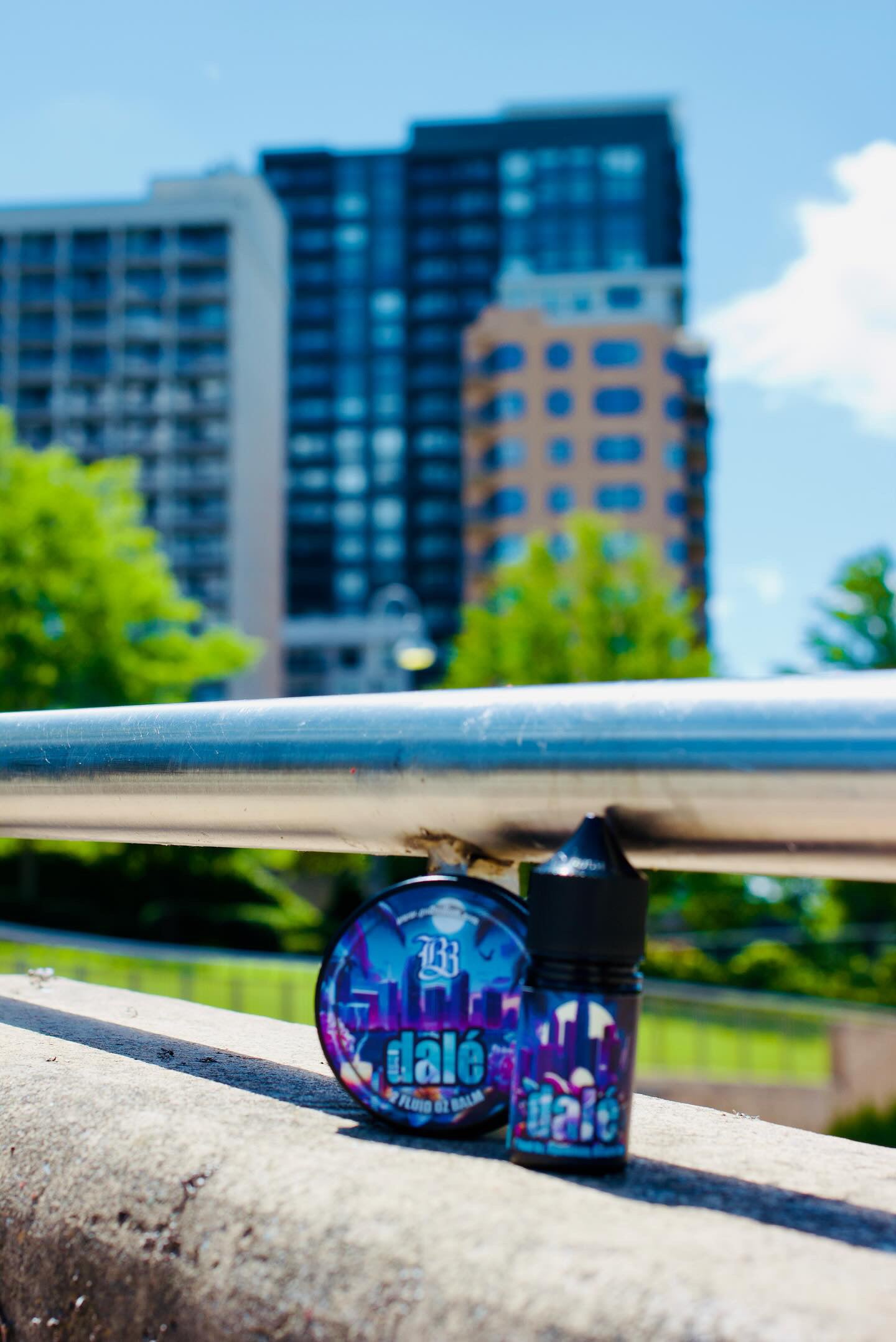 A round tin of Bedlam Beard Companys DALÉ Beard Balm and a small bottle of DALÉ Beard Oil, both with vibrant labels, sit on a concrete ledge with tall buildings and green trees blurred in the sunny background.