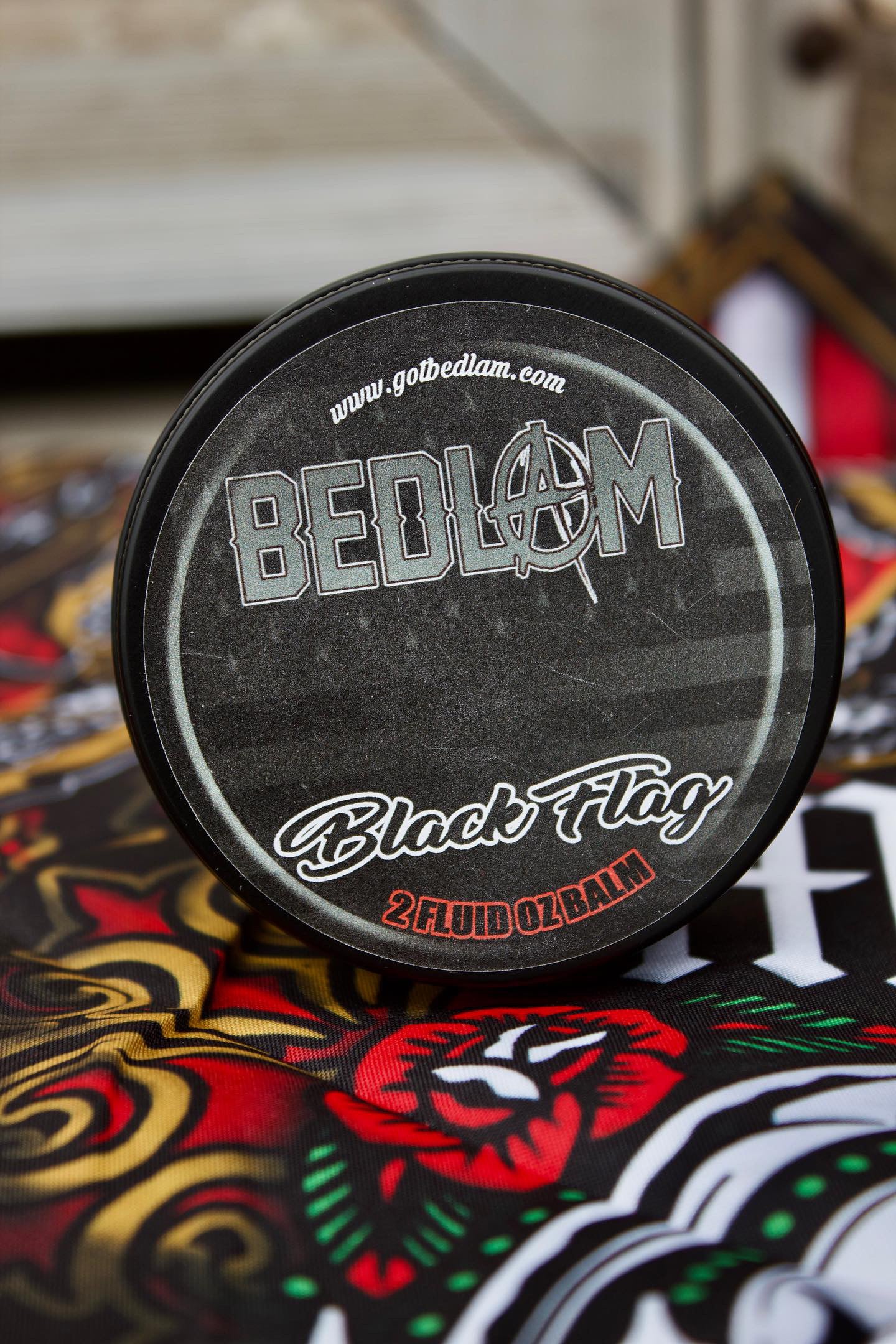 A round black container labeled Bedlam Beard Company BLACK FLAG Beard Oil & Balm sits on a colorful, tattoo-style surface. Perfect for fans of masculine scents, it features www.gotbedlam.com at the top of the lid.