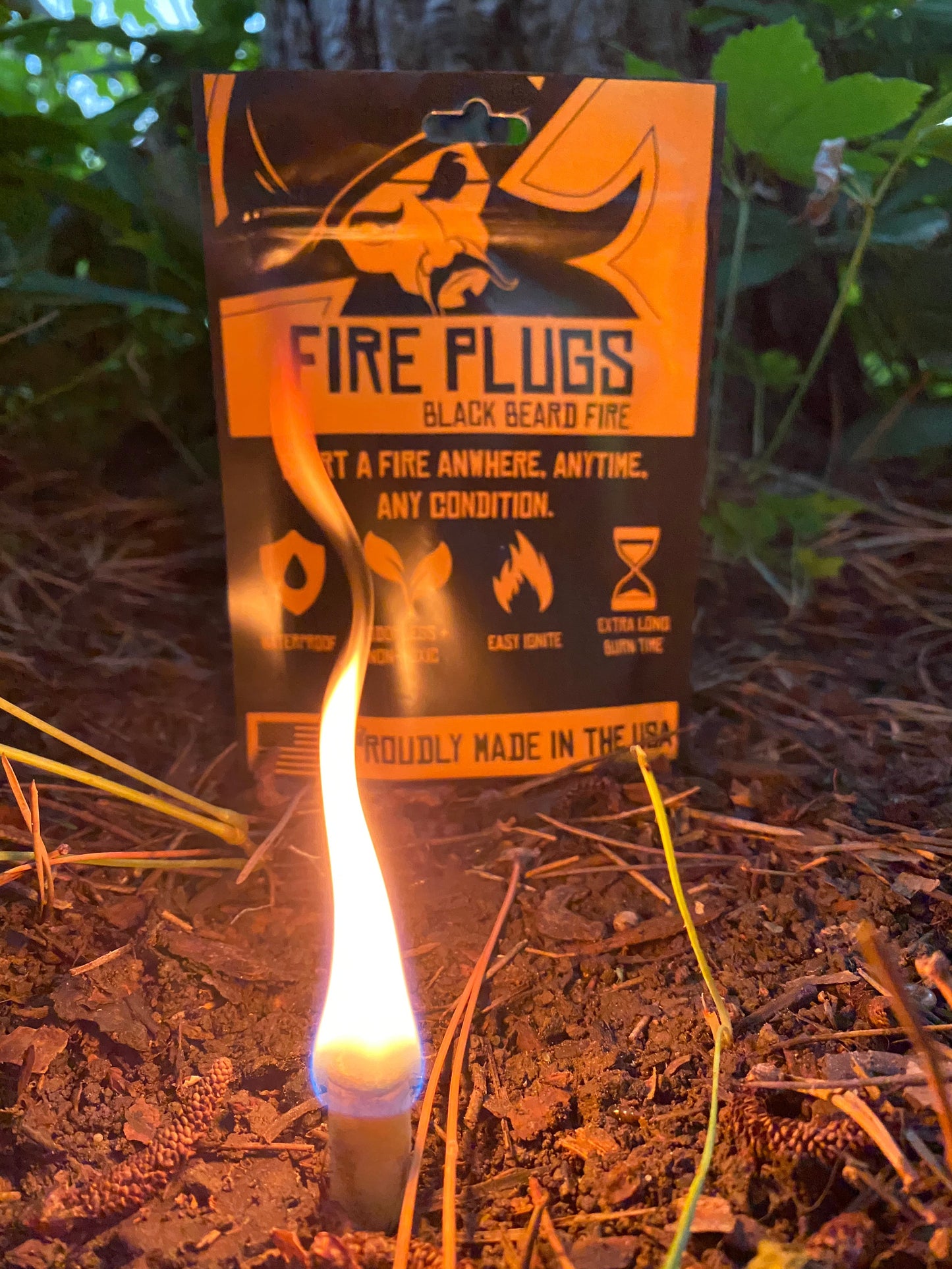 A lit Fire Plugs - Fire Starter Tinder by Black Beard Fire Starters burns near pine needles in front of its packaging, which shows icons for easy ignition and long burn time. Green foliage is seen in the background.