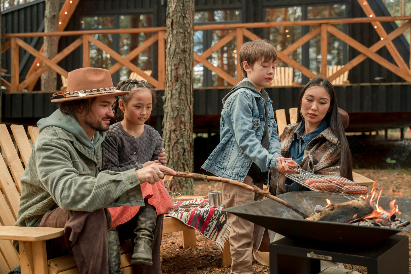 A family of four roasts sausages on skewers around an outdoor fire pit, using Black Beard Fire Starters’ Fire Plugs - Fire Starter Tinder. Behind them, a modern cabin with large windows and string lights nestles among the trees.