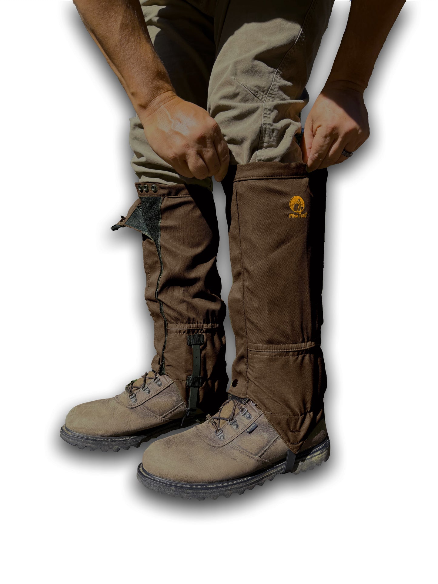 A person adjusts Pike Trail All-Season Waterproof Leg Gaiters—built for the trail, hunt, and more—worn over khaki pants and gray hiking boots. The gaiters display a small embroidered logo on the side.