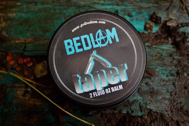 A round container labeled Bedlam Beard Company TAPER Beard Oil & Balm, featuring a straight razor graphic, sits on rustic turquoise and brown wood—hinting at a rich sandalwood bourbon scent among scattered objects and dirt.