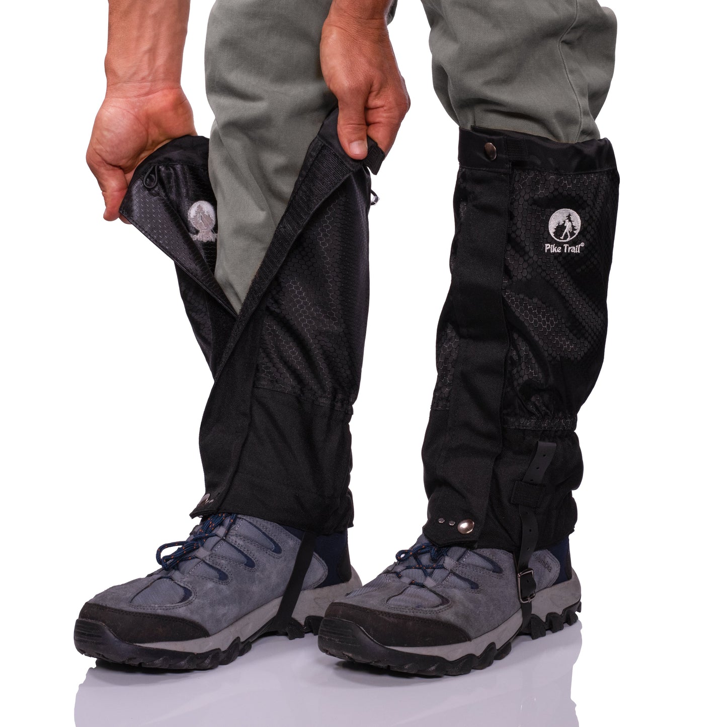 A person fastens Pike Trail All-Season Waterproof Leg Gaiters over gray hiking pants and blue boots, securing them with snaps and straps. The gaiters feature the Pike Trail logo and are designed for trail use, hunting, and outdoor activities.