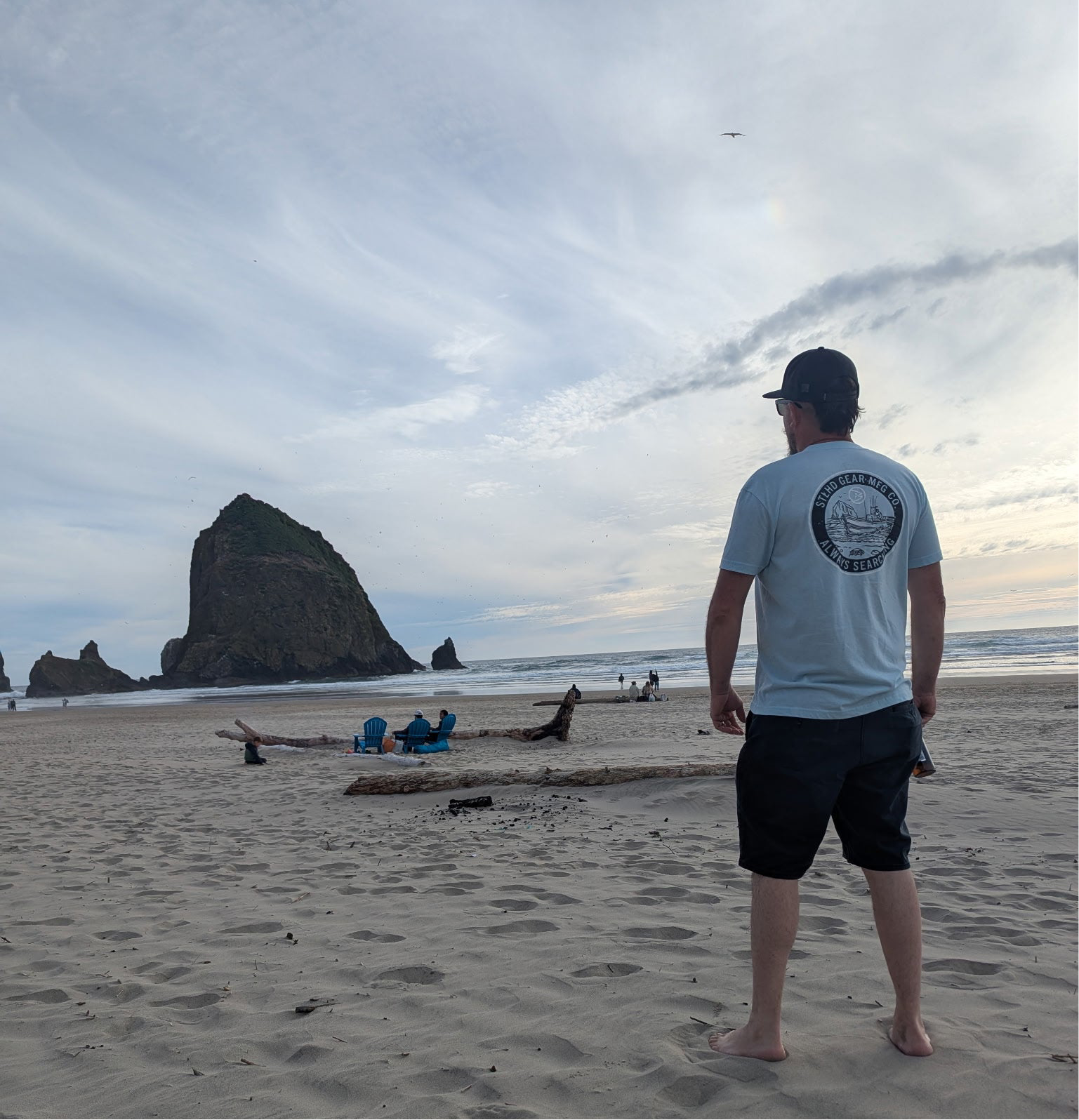 A barefoot man in a cap, shorts, and an STLHD GEAR Coastie T-Shirt stands on a sandy beach facing Haystack Rock, with driftwood and people in beach chairs under a cloudy sky.