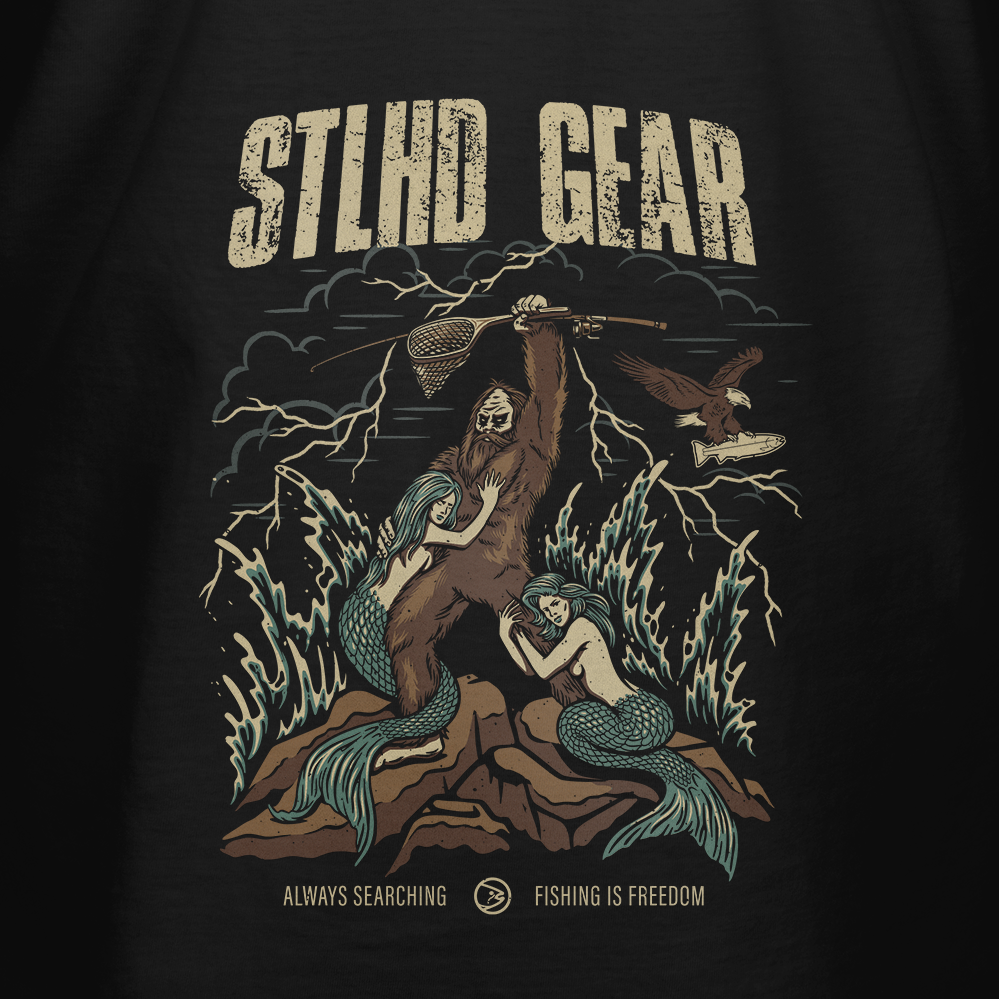 The STLHD King of the Mountain T-Shirt by STLHD GEAR features a hooded figure with a fishing net, flanked by two mermaids and an eagle, lightning in the background, and ALWAYS SEARCHING - FISHING IS FREEDOM—outdoor adventure vibes.