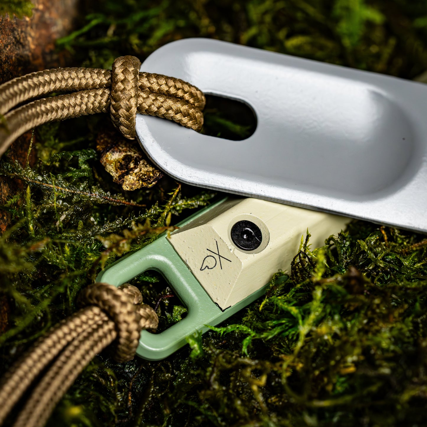 A close-up of the Überleben Hexå PRO, a compact multi-colored survival fire starter from Überleben, featuring a braided cord and metal tag, rests on green moss. Its engraved symbol and screw highlight its status as a key wilderness essential.