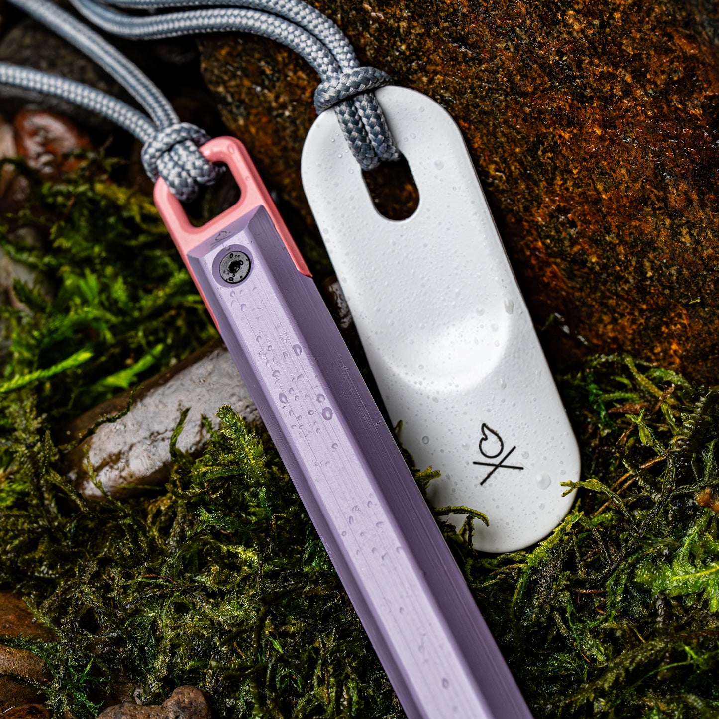 A close-up shows two pendants on cords resting on moss and rocks: one is a pastel purple stick with a pink end, and the other is the Überleben Hexå PRO – Refined Fire-Building Workhorse by Überleben, featuring a white, oval design with water droplets.