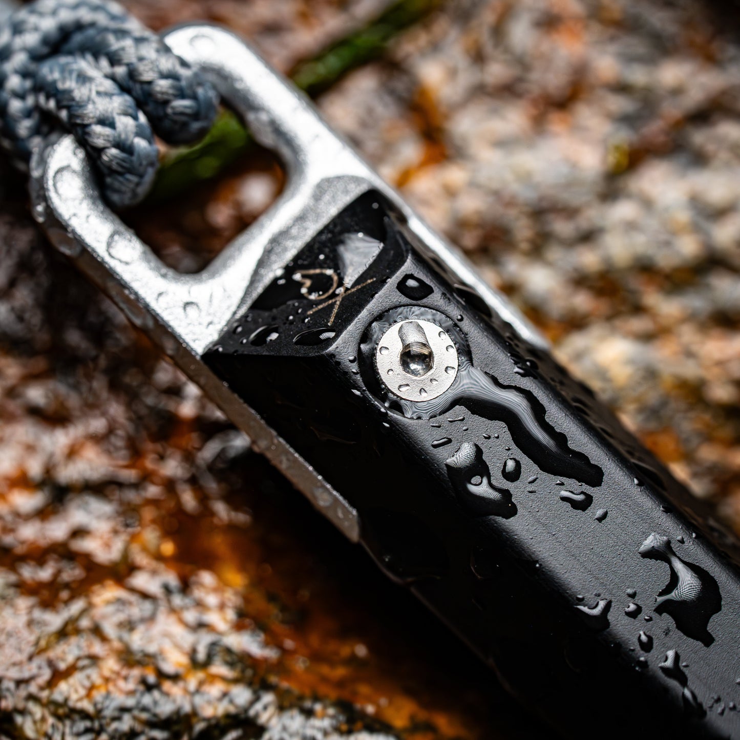 A close-up of the Überleben Hexå PRO by Überleben, a black multitool with water droplets on wet rock, featuring a metal bottle opener, gray cord, and integrated ferrocerium fire starter for survival situations.