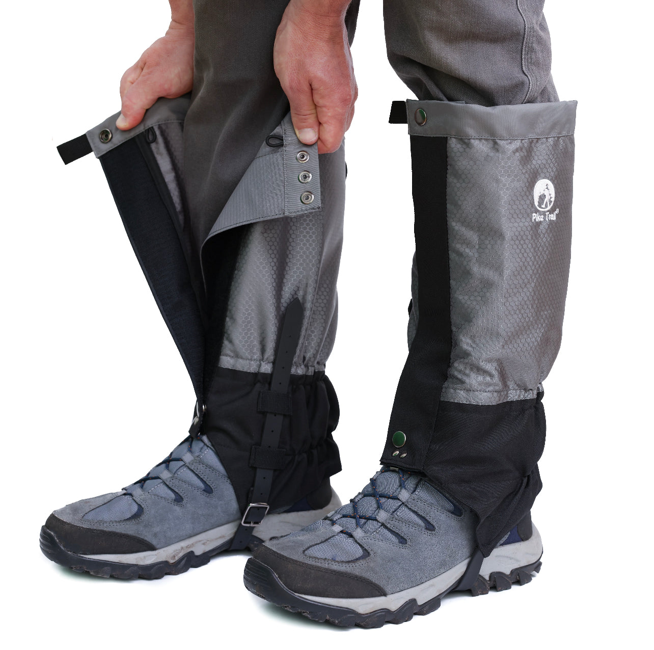 A person fastens Pike Trail All-Season Waterproof Leg Gaiters—gray and black, partially open to show snaps and Velcro—over boots and pants, getting ready for outdoor adventures.