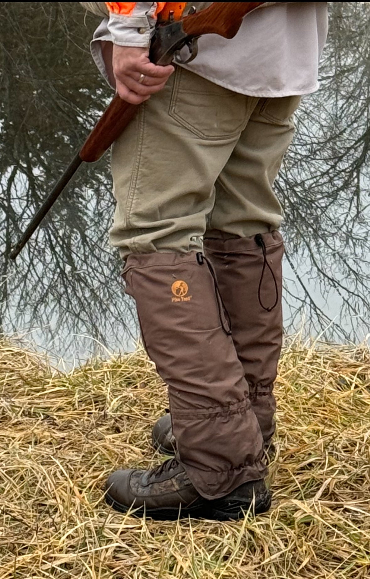 Wearing khaki pants, hiking boots, and Pike Trail All-Season Waterproof Leg Gaiters, a person stands on dry grass near water with a rifle. Bare tree branches are reflected in the background.
