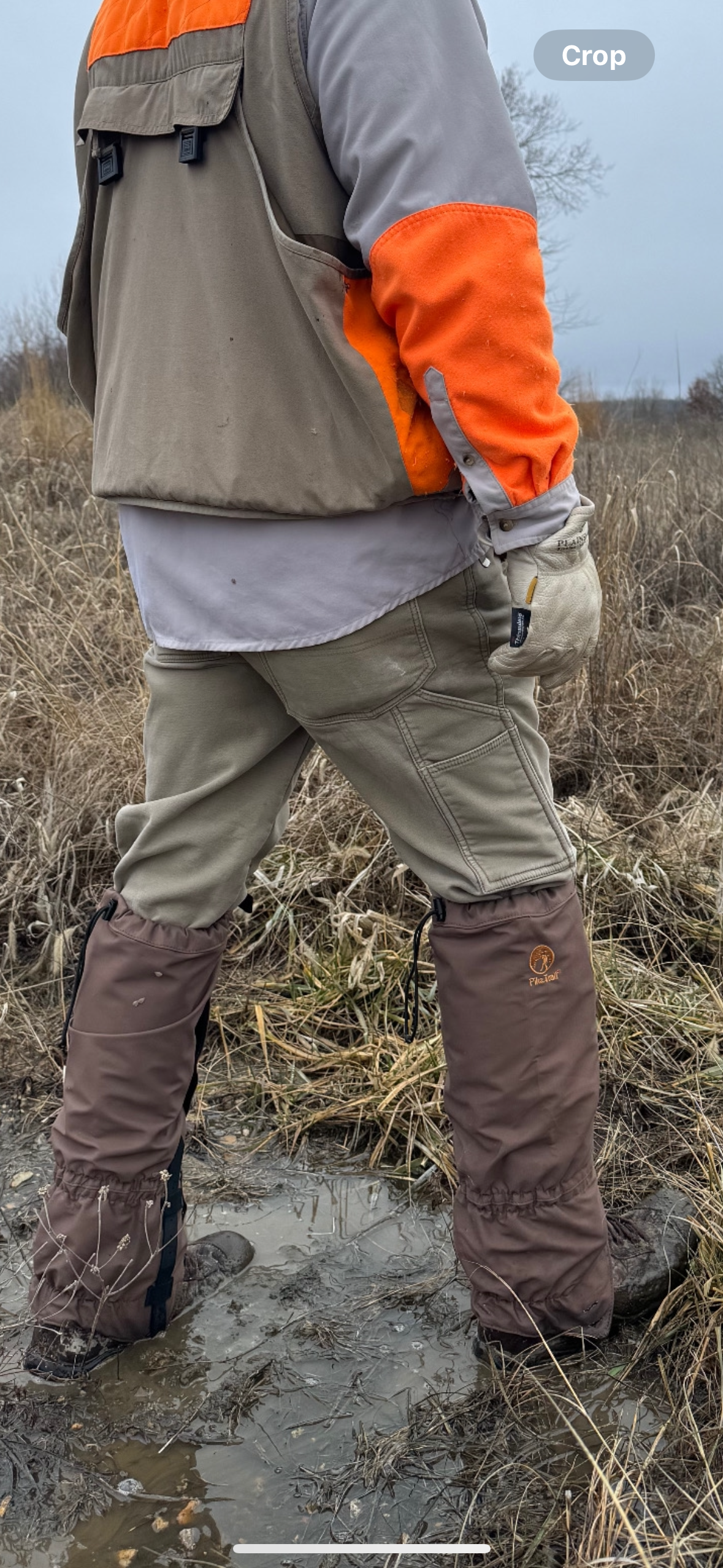 A person wearing Pike Trail All-Season Waterproof Leg Gaiters is seen from behind, standing in muddy grassland with tan pants and an orange jacket, showing the gaiters’ protection against wet and rugged terrain on a cloudy day.