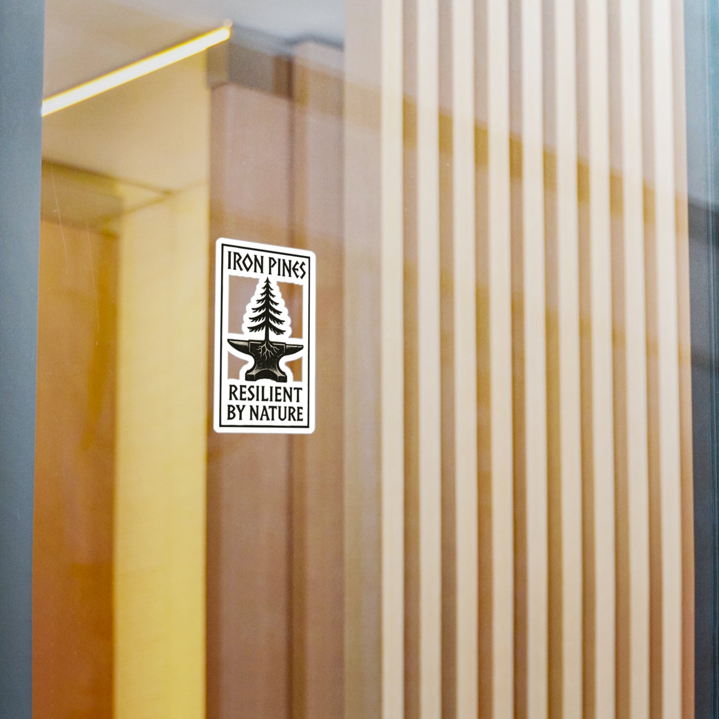 A durable vinyl Iron Pines Logo Sticker from Printify, featuring a pine tree with Iron Pines above and Resilient by Nature below, decorates a glass wall. Wooden slats and reflections in the background evoke resilience.