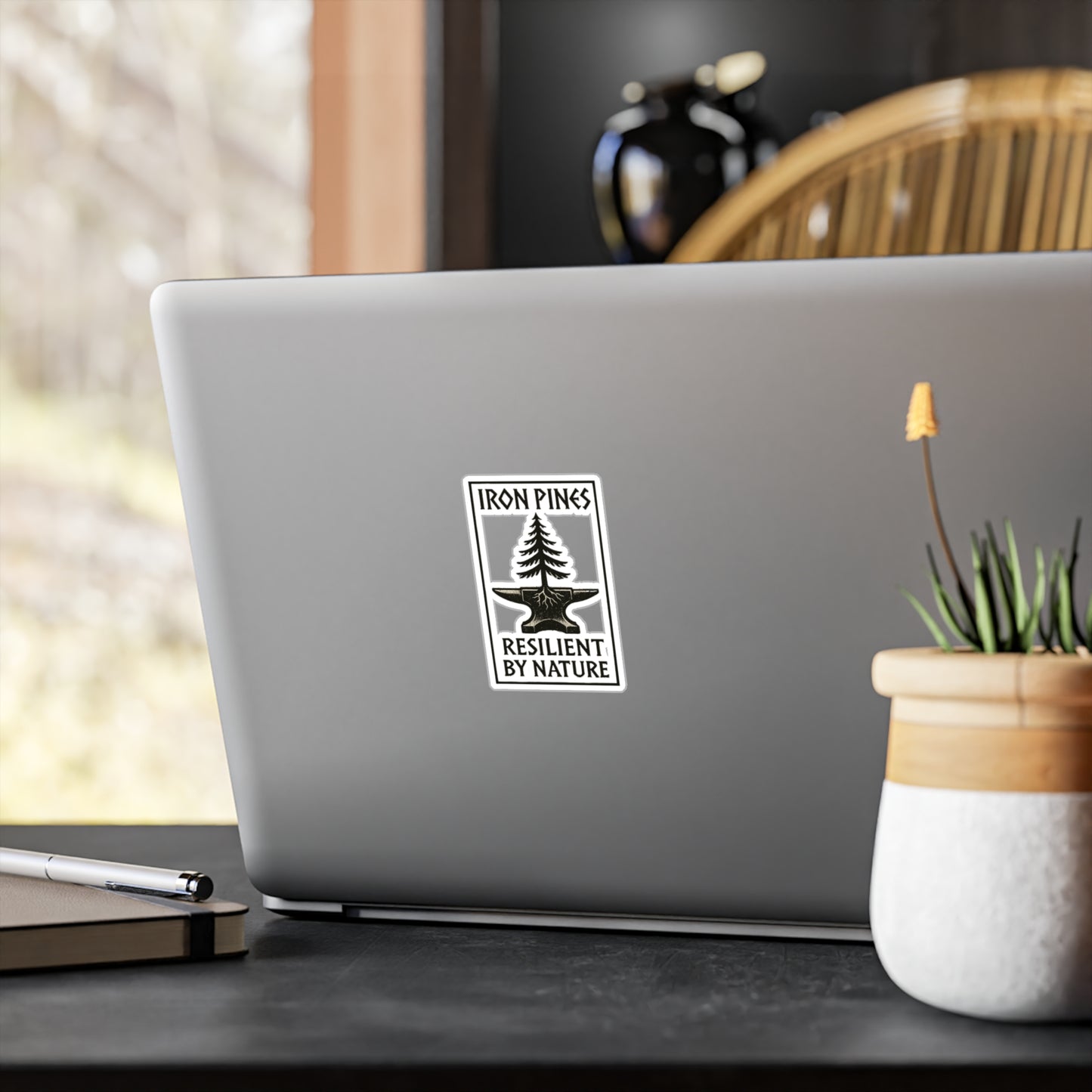 A silver laptop features the Iron Pines Logo Sticker – Durable Vinyl with Purpose by Printify, reading Resilient by Nature, on a desk beside a plant, pen, and notepad, inspiring resilience against a bright window view.