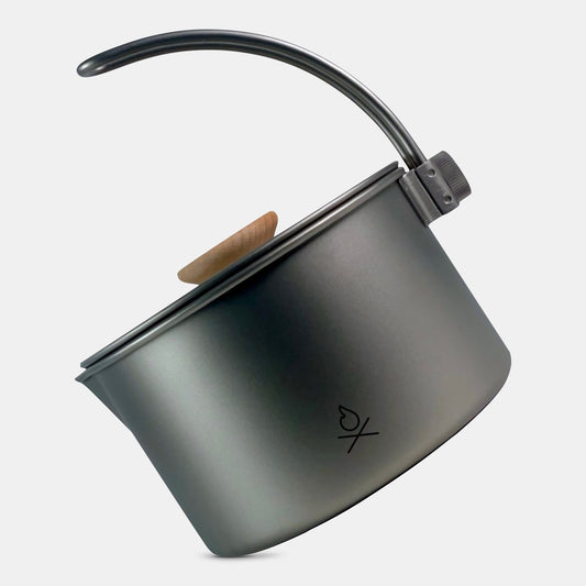 The Überleben Kessel – The Minimalist Cook Pot by Überleben is a gray metal titanium pot with a wooden lid knob, curved handle, and a small etched logo, shown against a light background. Designed for ultralight cooking needs.