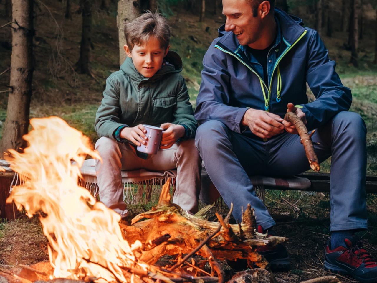 A man and a boy sit by a campfire in the woods, enjoying the warmth made possible by Black Beard Fire Starters Fire Plugs - Fire Starter Tinder. The boy holds a metal mug as they relax together in jackets amid the peaceful forest.