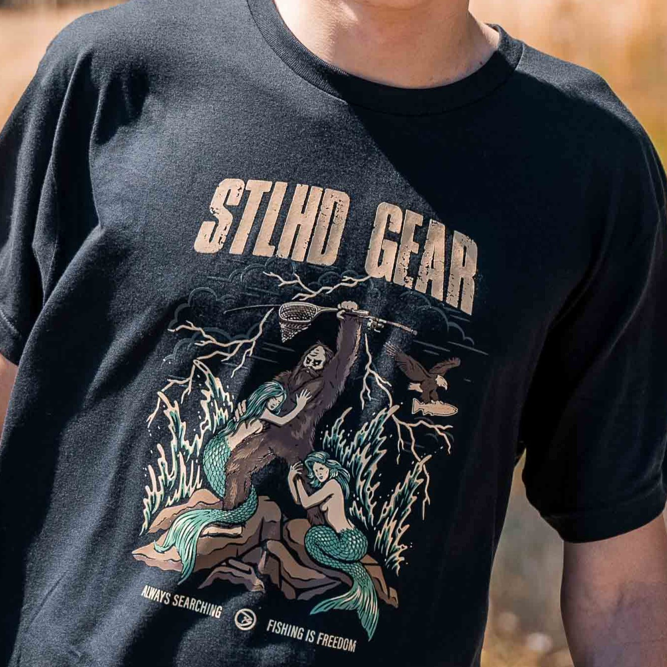A person wearing the STLHD King of the Mountain T-Shirt by STLHD GEAR, featuring an illustrated skeleton with a fishing rod, lightning, and two mermaids. The shirt reads: Always searching. Fishing is freedom.