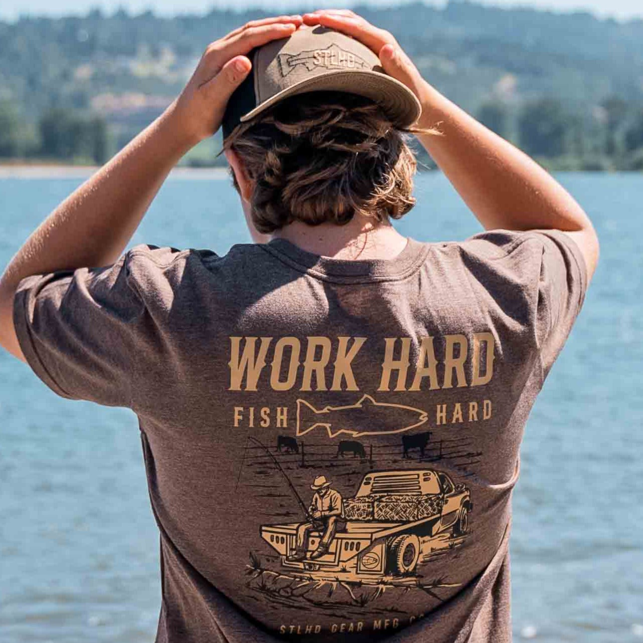 Wearing an STLHD GEAR Rancher T-Shirt that reads WORK HARD FISH HARD with fishing and truck art, a person in a cap stands by a lake, surrounded by trees and hills—capturing classic outdoor style.