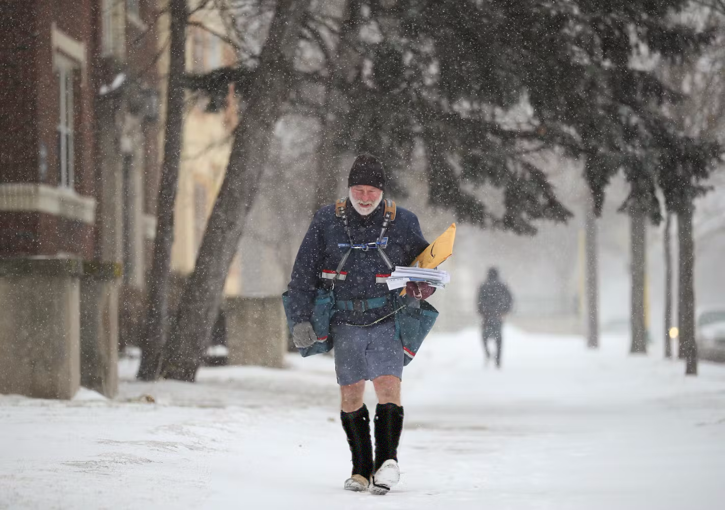 A postal worker wears Pike Trail All-Season Waterproof Leg Gaiters, shorts, a jacket, and long socks to deliver mail on a snowy winter street lined with trees and buildings as snow falls.