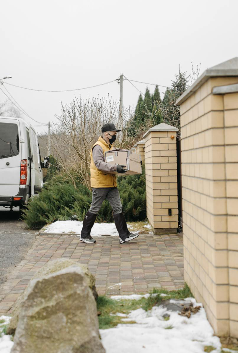 A delivery person in a yellow jacket and black hat, wearing Pike Trail All-Season Waterproof Leg Gaiters, carries a package toward a gated house on a snowy day, with a white van parked nearby and brick pillars at the entrance.