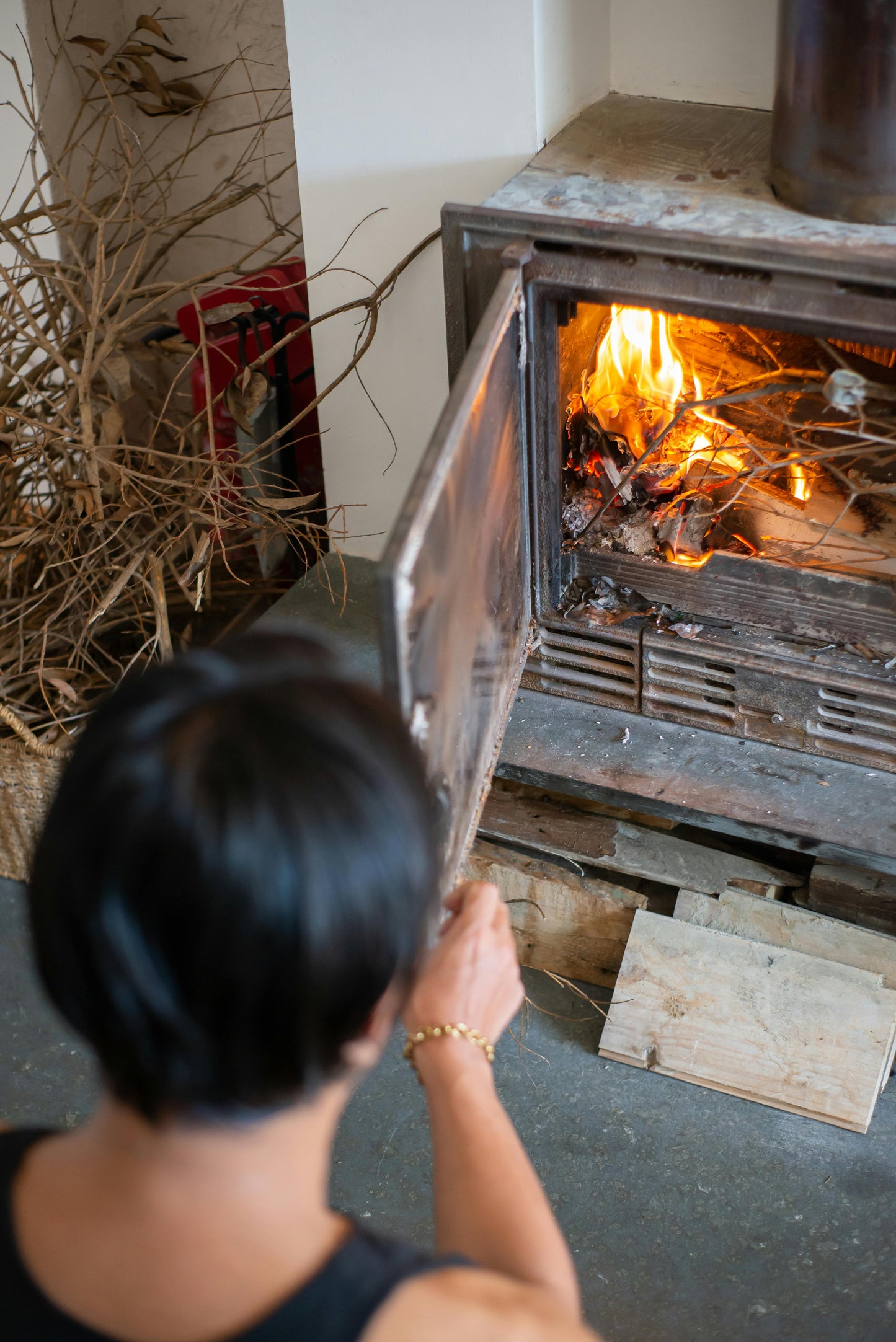 A person with short dark hair opens a wood-burning stove and adds kindling and Black Beard Fire Starters Fire Plugs - Fire Starter Tinder to a small indoor fire, while stacked branches are visible nearby.
